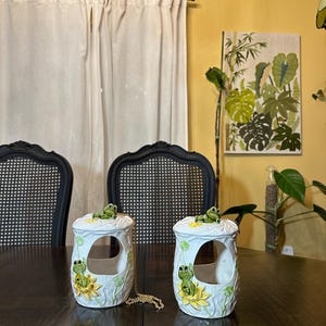 May include: Two white ceramic frog-themed containers with a gold chain. Each container has a frog on top and a frog on a sunflower. The containers are on a dark brown table in front of a yellow wall.