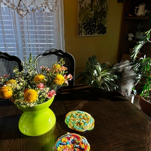 May include: A green vase filled with yellow and white flowers sits on a dark wood table with two colorful crocheted coasters.
