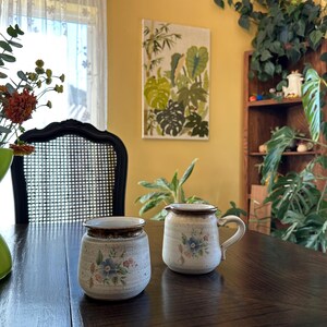 May include: Two ceramic creamers with floral designs on a dark wood table. The creamers are white with brown accents and blue and pink flowers.