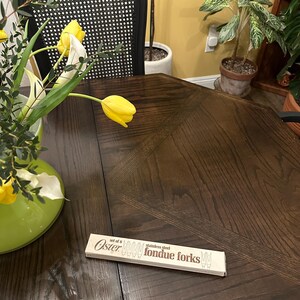 May include: A set of six stainless steel fondue forks in a brown box with the word "Oster" printed on it. The box is sitting on a dark brown wooden table with a bouquet of yellow flowers in a green vase.