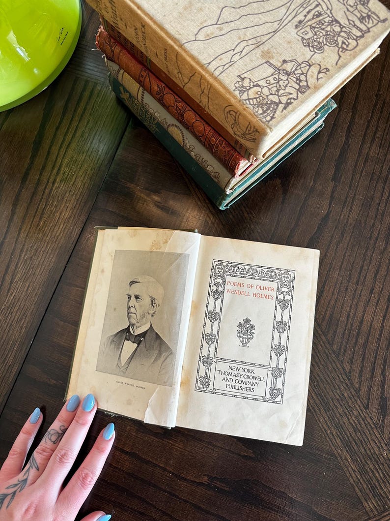 May include: An open book with a portrait of Oliver Wendell Holmes on the left page and the title "Poems of Oliver Wendell Holmes" on the right page. The book is published by Thomas Y. Crowell and Company, Publishers, New York.