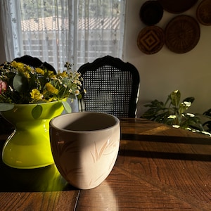 May include: A beige ceramic planter with a brown leaf design sits on a wooden table. The planter is empty and has a smooth, rounded shape.