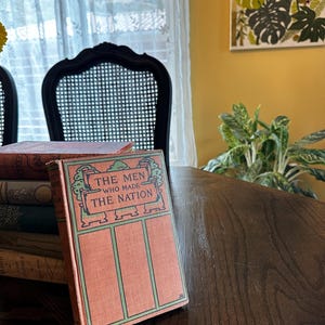 May include: A vintage book titled "The Men Who Made The Nation" with a green and orange cover. The book is sitting on a wooden table with other books stacked underneath.