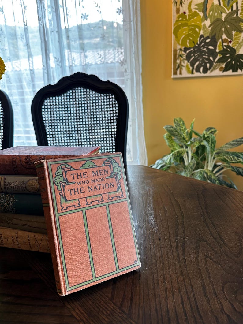 May include: A vintage book titled "The Men Who Made The Nation" with a green and orange cover. The book is sitting on a wooden table with other books stacked underneath.