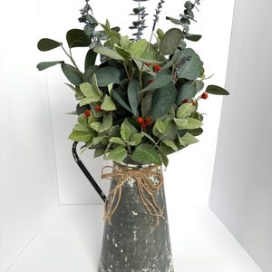 May include: A rustic, distressed metal pitcher filled with a variety of faux greenery. The arrangement includes eucalyptus, lavender, and small red berries. A twine bow adorns the pitcher's neck, adding a touch of farmhouse charm. The pitcher has a black handle.