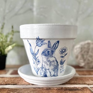 May include: White ceramic flower pot with a blue and white illustration of a rabbit and floral elements. The pot sits on a matching saucer. The design is reminiscent of classic blue and white china.