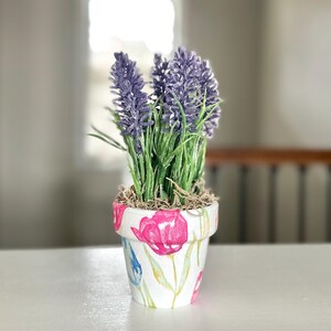 May include: A small, white ceramic flower pot with a floral design, holding artificial lavender. The pot features pink and blue tulip illustrations with green leaves. The lavender has purple flower spikes and green stems.