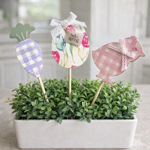 May include: Three Easter-themed garden stakes in a rectangular white planter filled with greenery. The stakes include a carrot, an egg, and a chick. The carrot has a purple and white checkered pattern. The egg has a floral design and a tag that says "hello spring". The chick has a pink and white checkered pattern.