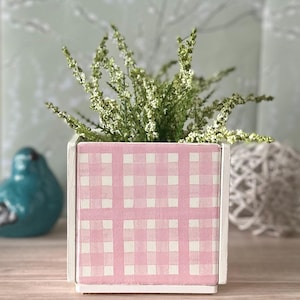 May include: A square, white-framed planter box with a pink and white gingham pattern. The box is filled with green and white flowering plants. A blue bird figurine is visible in the background.