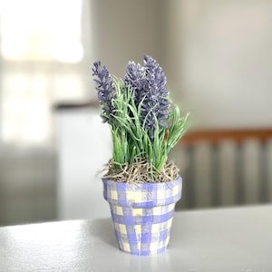 May include: A small potted lavender plant with purple flowers and green leaves. The pot is painted with a blue and yellow checkered pattern. The plant is a decorative item.