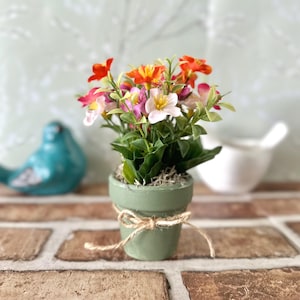 May include: A small, green ceramic flower pot tied with twine, filled with colorful artificial flowers. The flowers include orange, pink, and white blooms with green foliage. The pot sits on a brick surface, with a teal bird figurine in the background.