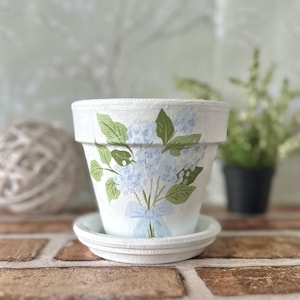 May include: White ceramic flower pot with a matching saucer. The pot is decorated with a blue and green floral design, featuring a light blue bow. The pot is sitting on a brick surface.