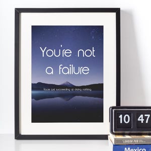May include: A framed print with the text "You're not a failure" against a night sky backdrop. The image features a mountain reflected in a lake. A clock and books are visible in the foreground.