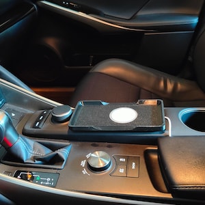 Puede incluir: Un interior de coche negro con una plataforma de carga inalámbrica para un teléfono. La plataforma está ubicada en la consola central y tiene un círculo blanco en el centro. El coche tiene un interior de cuero negro y una palanca de cambios plateada.