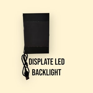 Pode incluir: Luz de fundo LED retangular preta com um cabo USB. O texto "DISPLATE LED BACKLIGHT" está impresso abaixo do dispositivo.