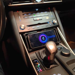 Puede incluir: El interior de un coche con un salpicadero de cuero negro y una palanca de cambios de plata. Un teléfono inteligente está montado en un soporte sobre la palanca de cambios. La pantalla del teléfono muestra una aplicación de navegación con un círculo azul y morado en el centro.