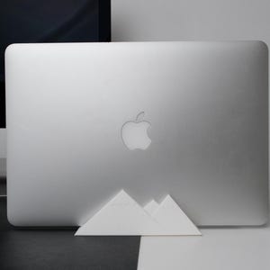 以下が含まれることがあります： 白いラップトップコンピューターは、シルバー仕上げで、アップルのロゴが付けられています。白い三角形のスタンドの上に置かれています。ラップトップは黒い表面の上にあります。