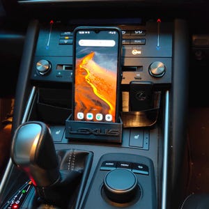 Pode incluir: Um interior de carro preto com um telefone montado em um suporte de telefone preto que diz "LEXUS". O telefone mostra uma imagem de paisagem de uma cena desértica. O carro tem uma alavanca de câmbio, um painel de controle com botões rotulados como "AUDIO" e "MENU", e um relógio digital que marca 12:59.