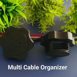 Pode incluir: Dois organizadores de cabos de plástico preto, um com a parte superior redonda e o outro com a parte superior estriada. O texto "Multi Cable Organizer" está impresso abaixo dos organizadores.