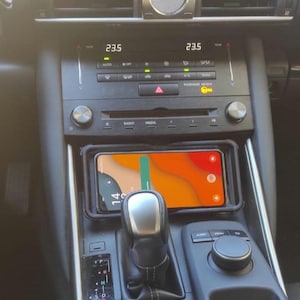 Puede incluir: Un interior de coche negro con una pantalla digital que muestra la hora como 23.5. El coche tiene una palanca de cambios y una plataforma de carga de teléfono con un teléfono en ella. La pantalla del teléfono muestra un mapa con un fondo verde y naranja.
