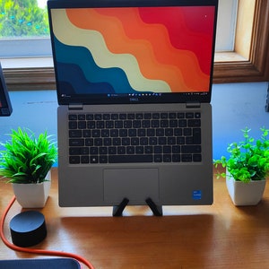 Pode incluir: Um laptop Dell prateado com um papel de parede abstrato colorido na tela está em um suporte de laptop preto. O laptop está em uma mesa de madeira com um mousepad preto e um cabo preto e laranja. Há duas pequenas plantas em vasos na mesa.