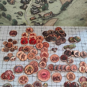 May include: A collection of fantasy role-playing game tokens featuring various creatures and characters. The tokens are circular with detailed illustrations and are scattered on a gridded game mat. A map of a village is in the background.