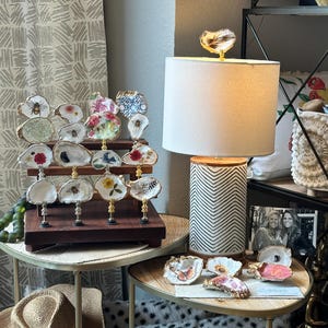 May include: A display of decorative oyster shell art with various designs, including flowers, bees, and abstract patterns. The shells are mounted on gold-toned stands and arranged on a dark wooden display. A lamp with a cream shade and a patterned base is also visible.
