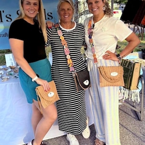 May include: Three women standing together, each holding a small, woven straw purse with a flap closure. The purses have a natural tan color and are adorned with fringe. The women are wearing casual clothing, including a black t-shirt, a striped dress, and striped pants.