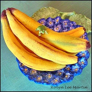 Può includere: Un mazzo di banane gialle su un piatto floreale blu e bianco con una statuetta di rana verde in cima.