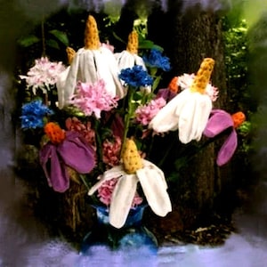 以下が含まれることがあります： 白、ピンク、青の花束で、中心は黄色です。花は花瓶に活けられ、緑の葉で囲まれています。