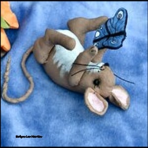 Pode incluir: Uma figurinha de rato marrom e branco com uma cauda longa, segurando uma borboleta azul em suas patas. O rato tem um nariz rosa e orelhas grandes. A figurinha está sobre um fundo de tecido azul.