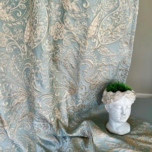 May include: A blue fabric with a gold paisley pattern. The fabric is draped over a surface and a decorative bust is in the background.
