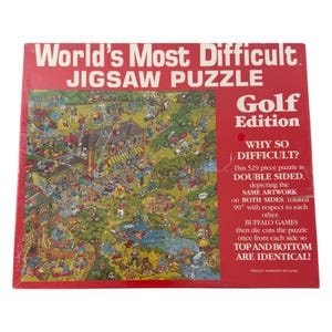 Könnte beinhalten: Ein quadratischer Puzzlekarton mit rotem Rand und weißem Text World's Most Difficult Jigsaw Puzzle Golf Edition. In der Mitte befindet sich eine bunte Cartoon-Illustration eines Golfplatzes. Der Karton gibt an, dass es ein 529-Teile-Puzzle ist.