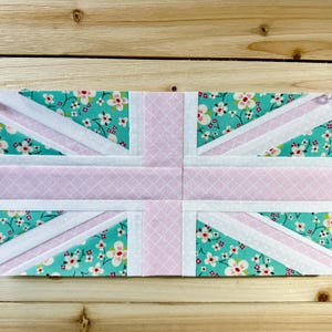 May include: A fabric Union Jack flag design. The flag features a light pink and white cross, with turquoise sections adorned with a floral pattern. The flag is held in place with clear pins.