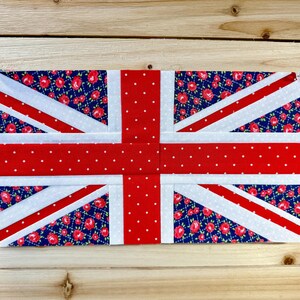 May include: A fabric Union Jack flag with a red and white design. The flag features a red cross and diagonal stripes on a blue floral background. The flag is mounted on a wooden surface.