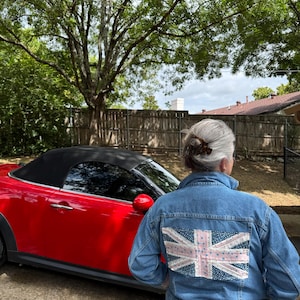 May include: A denim jacket with a Union Jack design on the back. The jacket is a medium wash blue and is paired with matching jeans. A red convertible car is parked nearby, with a black top.