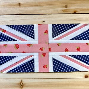 May include: A rectangular fabric item resembling the Union Jack flag. The design incorporates white, red, and navy blue. The navy blue sections feature white diagonal stripes. The red sections are adorned with small strawberry patterns. The item is displayed on a wooden surface.