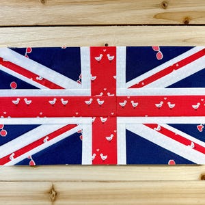 May include: A rectangular fabric item featuring a Union Jack design. The design incorporates red, white, and blue colors, with white polka dots and small bird illustrations. The item is held together with clear pins.