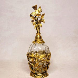 May include: A clear glass perfume bottle with a gold-colored bird and floral design. The bottle has a round shape with vertical ridges and is adorned with gold-colored leaves and flowers. The bird sits atop the bottle's stopper.
