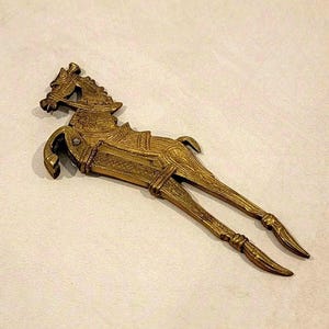 May include: A brass-colored metal nutcracker shaped like a stylized horse in a leaping position. The object features intricate engraved patterns along the body, head, and legs, with a hinged mechanism at the front legs for cracking nuts.