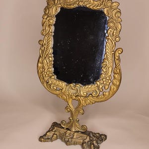 May include: Ornate gold-colored vanity mirror with a rectangular black mirror. The frame features intricate floral and scrollwork designs. The mirror is mounted on a decorative base, adding to its vintage aesthetic.