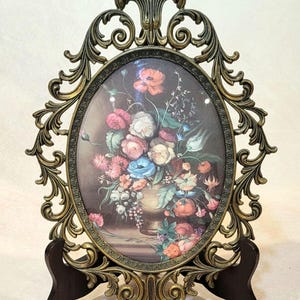May include: An oval-shaped picture frame with an ornate, antique brass-colored metal border featuring intricate scrollwork. The frame holds a print of a colorful floral bouquet in a vase, displaying red, white, pink, and blue flowers on a dark background.