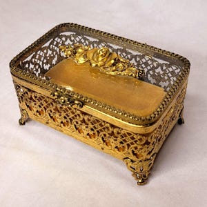 May include: A rectangular, gold-toned jewelry box with a glass top and ornate filigree detailing. The box features a decorative rose motif and rests on four small, decorative feet. Inside, a gold-colored cushion provides a soft base.