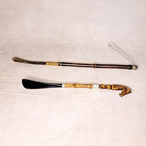 May include: Two shoe horns. One is dark brown with a gold-colored metal tip and a gold-colored coil near the handle. The other is black with a bamboo handle and a gold-colored metal accent, and a decorative end.