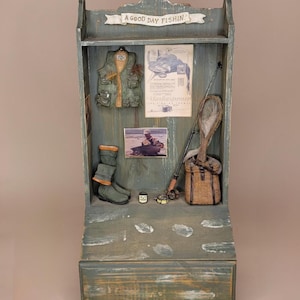 May include: A wooden display shelf with fishing-themed decor. It features a vest, boots, a fishing rod, a net, and a basket. The shelf has the text "A GOOD DAY FISHIN!" and a vintage advertisement.