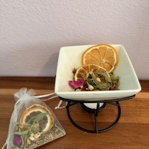 May include: A white ceramic essential oil diffuser with a black metal stand, filled with dried herbs and orange slices. A small organza bag filled with similar botanicals sits to the left. The items are on a wooden surface.