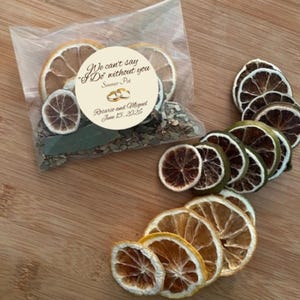 May include: A clear cellophane bag filled with dried herbs and citrus slices, with a circular sticker that reads "We can't say 'I Do' without you". Several dried citrus slices are arranged around the bag on a wooden surface.