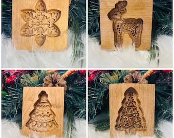 Könnte beinhalten: Vier hölzerne Keksausstecher mit festlichen Designs. Die Formen zeigen eine Schneeflocke, ein Rentier und zwei Weihnachtsbäume. Die Designs sind in das Holz geschnitzt und zeigen komplizierte Details. Die Formen werden auf einer weißen, flauschigen Oberfläche mit Weihnachtsgrün präsentiert.