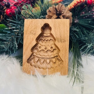 May include: A wooden cookie mold in the shape of a Christmas tree. The mold is rectangular with a detailed tree design carved into the wood. The tree has multiple layers with decorative elements. The wood has a light brown color.