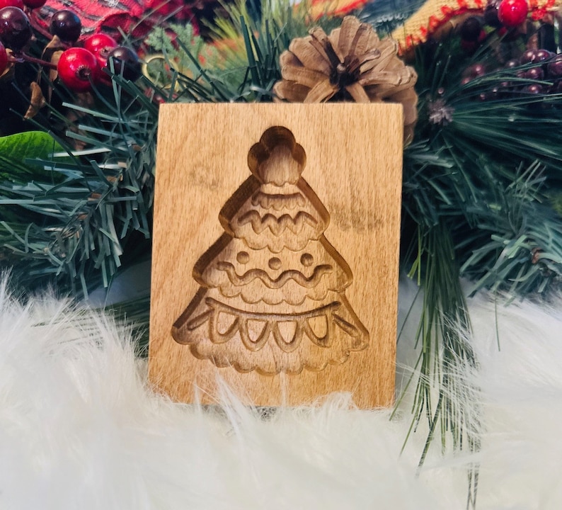 May include: A wooden cookie mold in the shape of a Christmas tree. The mold is rectangular with a detailed tree design carved into the wood. The tree has multiple layers with decorative elements. The wood has a light brown color.